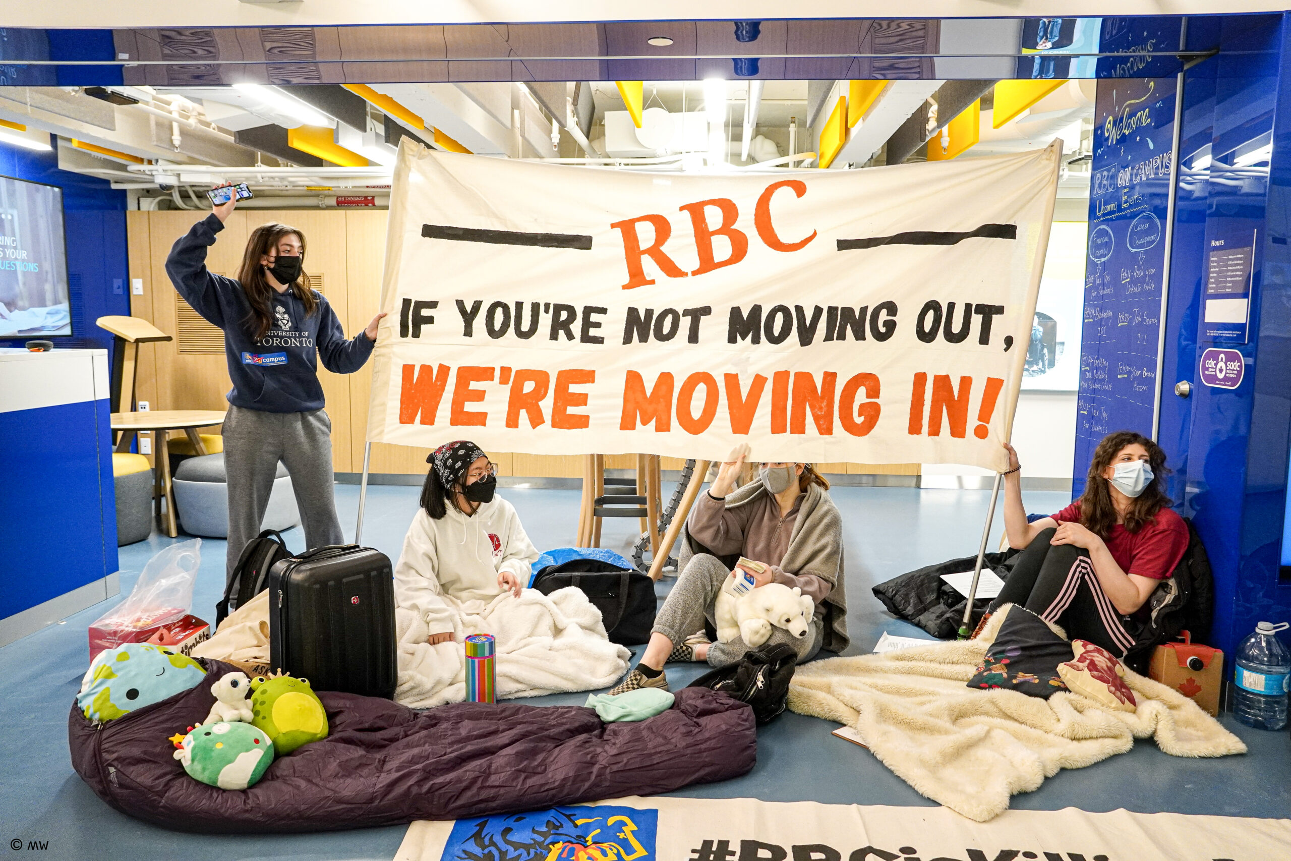 A photo of Climate Justice University of Toronto students doing a sit-in protest in the RBC OnCampus branch. A white woman in a red shirt sits on the ground holding the base of a banner. Another white person is partially visible behind the banner, a chinese woman sits in front, they are all on blankets and pillows. A green pillow pet stuffed animal is visible. A white woman with brown hear wearing a sweat suit stands beside the banner with her fist raised. The banner reads "RBC, If you're not moving out, we're moving in!" All of the students are wearing masks.
