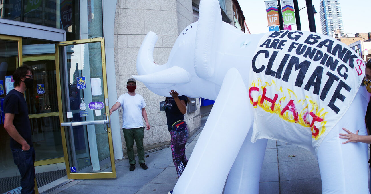 Photo of protesters attempting to bring a large white elephant inflatable into an RBC branch. Two people hold the door open while two people carry the inflatable. A banner is draped over the elephant that reads "Big banks are funding climate change."