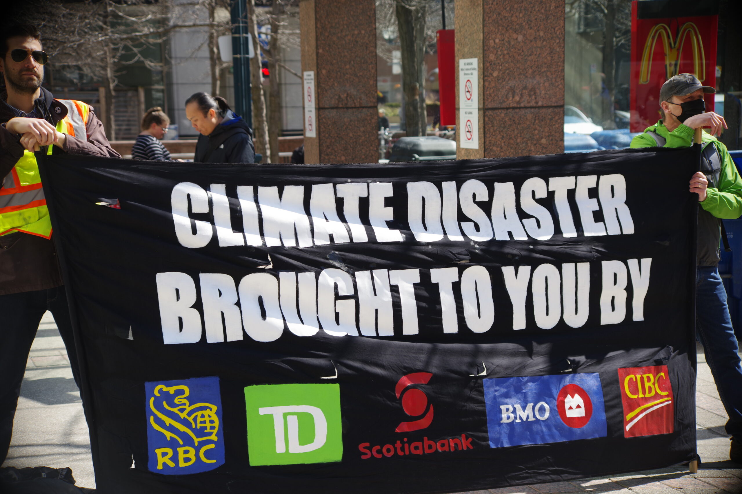 Two white men in yellow reflective safety vests are standing in a downtown Toronto street holding a black banner that reads "Climate disaster brought to you by" and then shows illustrations of the RBC, TD, Scotiabank, BMO and CIBC logos.