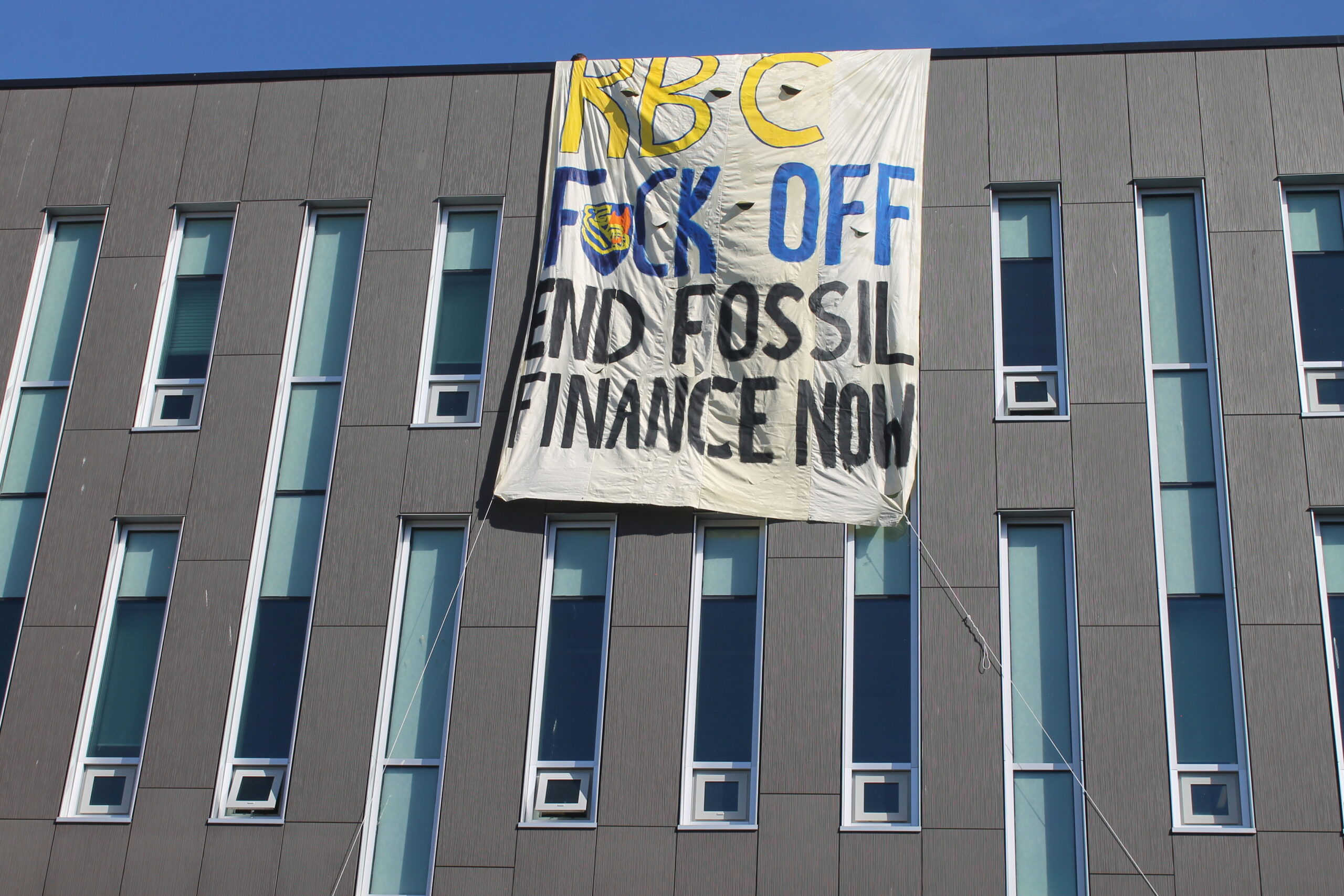 A grey university building with many long narrow windows. A strip of blue sky above. A large white banner hands over the roof covering more than a storey of the building. The banner reads "RBC FUCK OFF END FOSSIL FINANCE NOW."